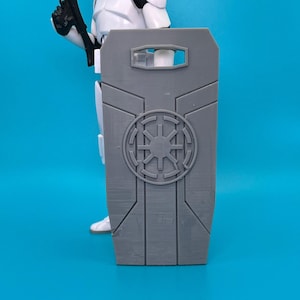 May include: A gray plastic shield with a raised Imperial symbol and a rectangular opening at the top. The shield is designed for action figures.