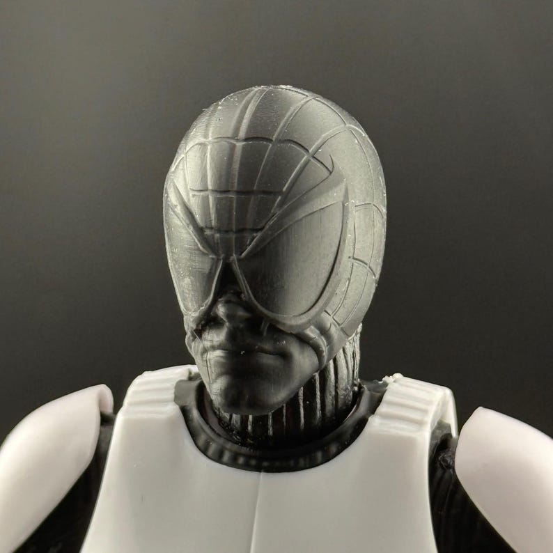 May include: A close-up of a black and white action figure's head and upper torso. The figure has a textured, web-like design on the helmet and large, stylized eye coverings. The figure is wearing a white chest piece.