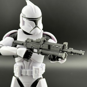 May include: A white action figure of a soldier in a helmet and armor, holding a gray rifle with a grenade launcher attachment. The figure has black accents on the helmet and gloves. The background is a solid dark gray.
