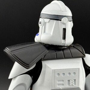 May include: A white and black action figure of a clone trooper, featuring a helmet with a dark gray visor and accents. The figure has black shoulder armor and a detailed chest plate. The overall design is reminiscent of a science fiction character.