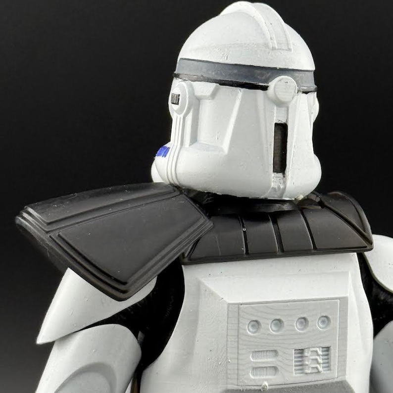 May include: A white and black action figure of a clone trooper, featuring a helmet with a dark gray visor and accents. The figure has black shoulder armor and a detailed chest plate. The overall design is reminiscent of a science fiction character.