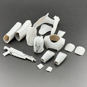 Peut inclure: Ensemble de pièces de modèle en plastique gris clair non assemblées, comprenant un casque, une pièce de poitrine, une armure de bras et de jambes, et un fusil blaster. Les pièces sont éparpillées sur une surface grise, suggérant un kit de modèle ou un projet de bricolage.