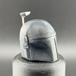 Pu&ograve; includere: Un casco grigio stampato in 3D con una piccola antenna sulla cima. Il casco ha una superficie testurizzata e si trova su una base cilindrica grigia.
