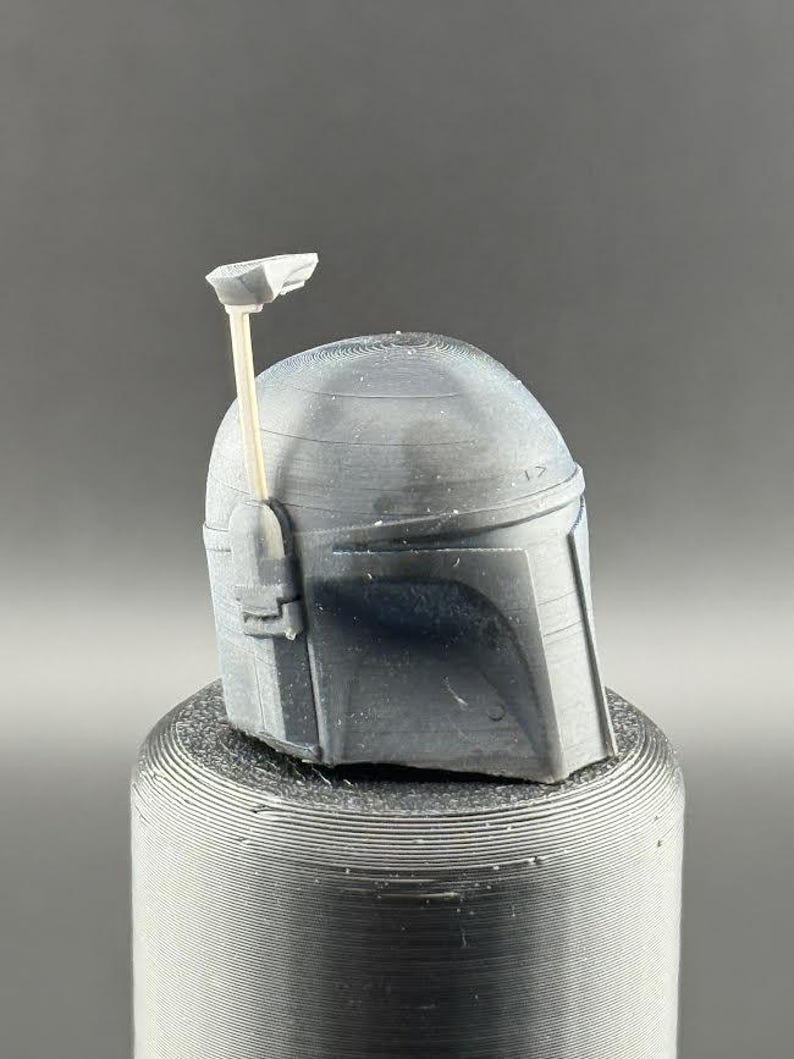 Pu&ograve; includere: Un casco grigio stampato in 3D con una piccola antenna sulla cima. Il casco ha una superficie testurizzata e si trova su una base cilindrica grigia.
