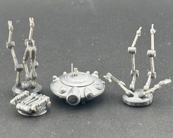 May include: Three miniature, gray, sci-fi spacecraft models with articulated arms. The spacecraft are on a gray surface.