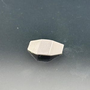 Pu&ograve; includere: Un piccolo oggetto geometrico grigio chiaro con un design sfaccettato. L'oggetto &egrave; posto su una superficie scura e riflettente, creando un effetto a specchio. La forma ricorda una gemma o un elemento architettonico stilizzato.