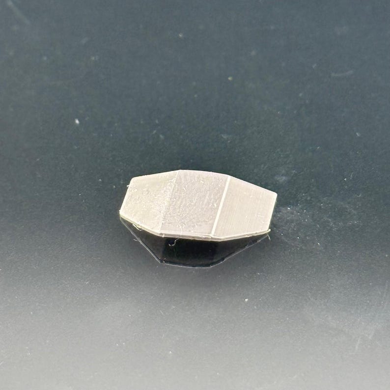 Pu&ograve; includere: Un piccolo oggetto geometrico grigio chiaro con un design sfaccettato. L'oggetto &egrave; posto su una superficie scura e riflettente, creando un effetto a specchio. La forma ricorda una gemma o un elemento architettonico stilizzato.