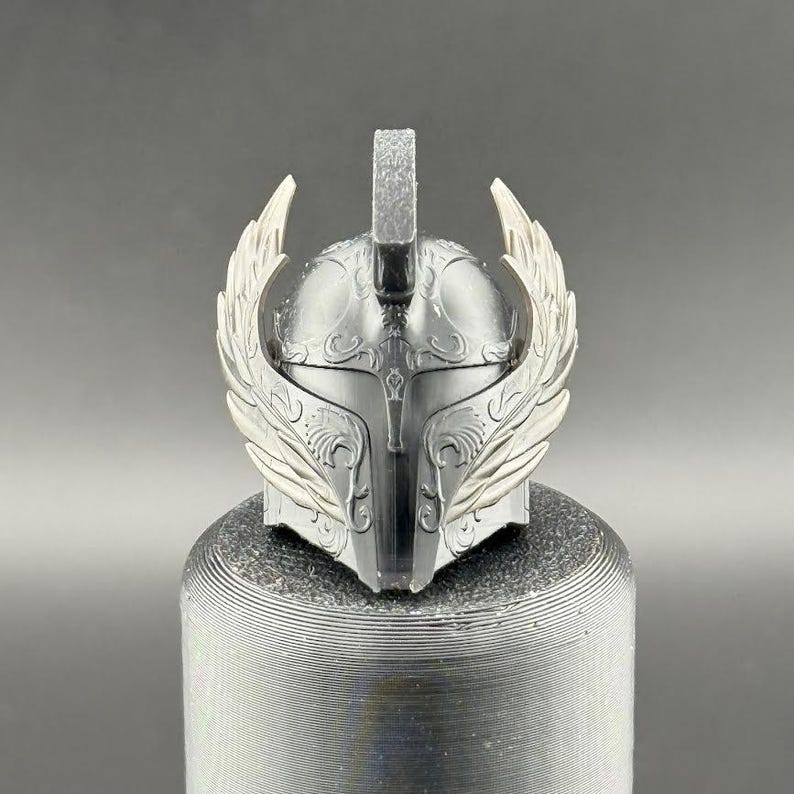 May include: A silver helmet with intricate details and a winged design. The helmet has a pointed top and a face mask with a decorative pattern.