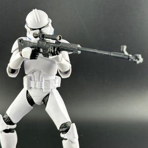 以下が含まれることがあります： ヘルメットと鎧を着た兵士の白いアクションフィギュア。黒いスナイパーライフルとスコープを持っています。フィギュアは射撃姿勢で、ライフルは前方を向いています。フィギュアは黒い手袋と白いベルトをしています。