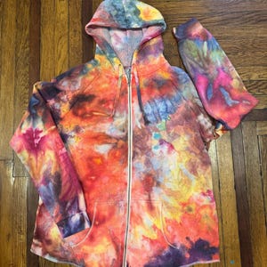 May include: A tie-dye zip-up hoodie in a vibrant mix of colors including orange, yellow, blue, and purple. The hoodie features a drawstring hood, a full-length zipper, and two front pockets. The sleeves are long and the fabric appears soft.