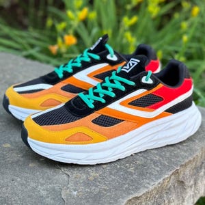 May include: A pair of athletic shoes with a black mesh upper, accented with orange, yellow, white, and red panels. The shoes have white soles and teal laces. The shoes are sitting on a gray stone.