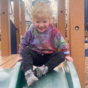 May include: A young child wearing a tie-dye hoodie and black pants sits on a green slide at a playground. The hoodie features a mix of purple, pink, and green colors. The child is smiling and wearing gray sneakers.