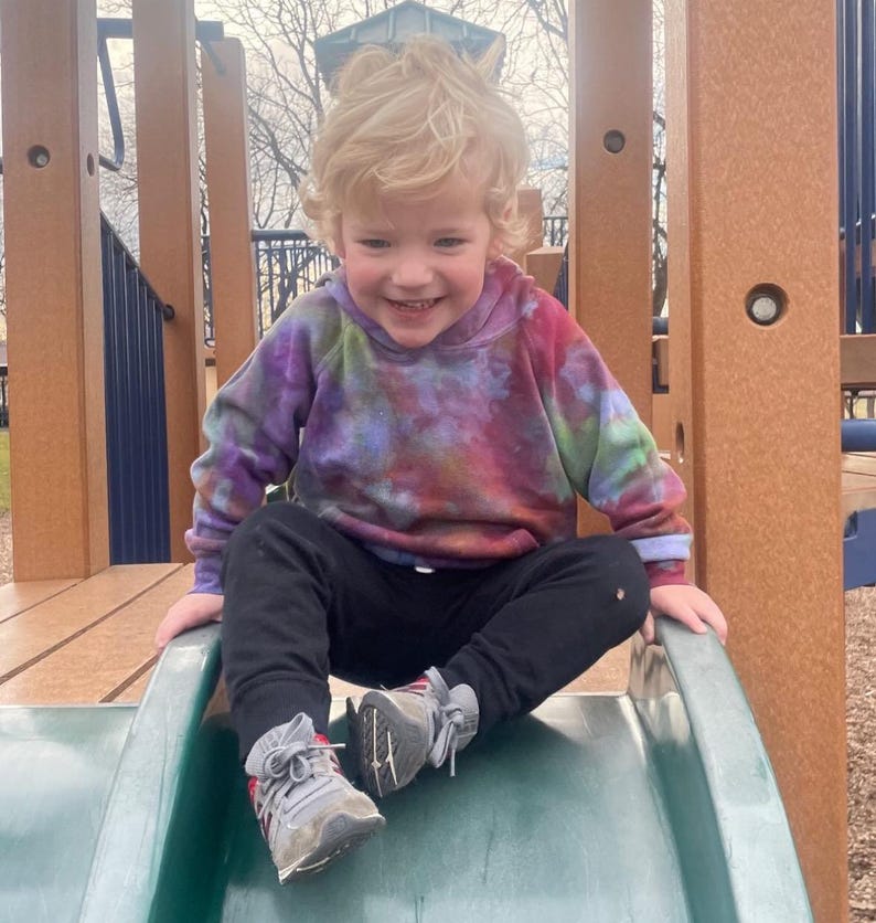 May include: A young child wearing a tie-dye hoodie and black pants sits on a green slide at a playground. The hoodie features a mix of purple, pink, and green colors. The child is smiling and wearing gray sneakers.