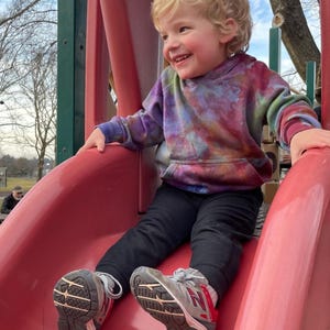 May include: A child wearing a tie-dye hoodie and black pants slides down a red playground slide. The child is smiling and wearing gray and red sneakers. The background includes trees and a park setting.