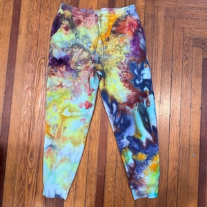 May include: Multi-coloured tie-dye sweatpants with a drawstring waist and pockets. The design features a blend of yellow, blue, purple, and red hues, creating an abstract pattern. The trousers are a comfortable fit.