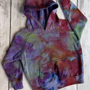 May include: A tie-dye hoodie in shades of purple, red, green, and blue. The hooded sweatshirt has a front pocket and ribbed cuffs. The garment is made of a soft material.