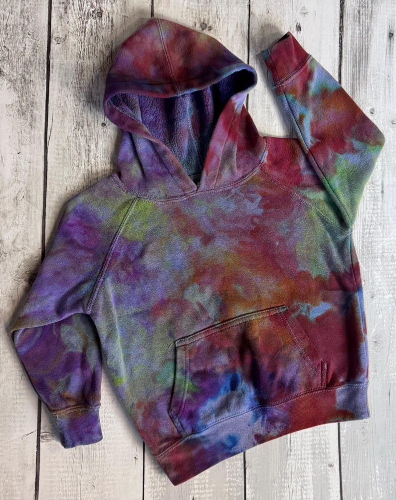 May include: A tie-dye hoodie in shades of purple, red, green, and blue. The hooded sweatshirt has a front pocket and ribbed cuffs. The garment is made of a soft material.