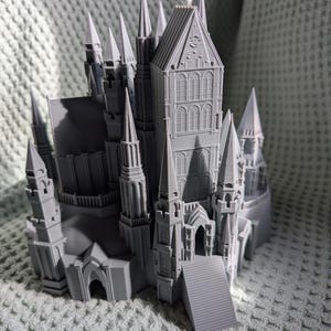May include: A gray, 3D-printed model of a castle with multiple spires, towers, and architectural details. The castle features arched windows, a central tower, and a staircase leading to an arched entrance. The model is set against a textured background.