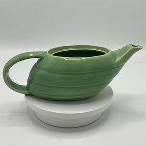 May include: A green ceramic teapot with a rounded body and a curved spout. The teapot has a smooth, glossy finish and is decorated with subtle horizontal lines.