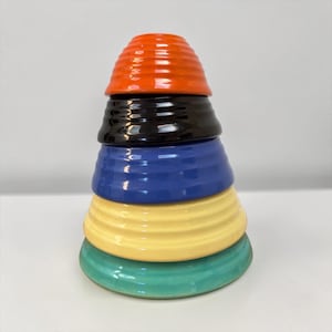 May include: A stack of five circular ceramic bowls with a ribbed texture. The bowls are arranged in descending order of size from top to bottom, featuring a vibrant color palette of orange, black, blue, pale yellow, and teal green.