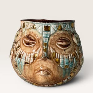 May include: A ceramic vessel featuring a sculpted face with closed eyes and a mouth. The surface is decorated with a mosaic-like pattern of light blue and brown glazes, accented with small, reflective gold-toned square tiles across the exterior.