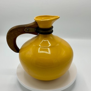 May include: A yellow ceramic pitcher with a wooden handle and a metal band around the neck. The pitcher is sitting on a white pedestal.