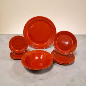 May include: A set of orange ceramic dinnerware featuring a large dinner plate, a deep bowl, and several smaller plates and saucers. Each piece is characterized by a glossy finish and concentric ring patterns along the outer rims.