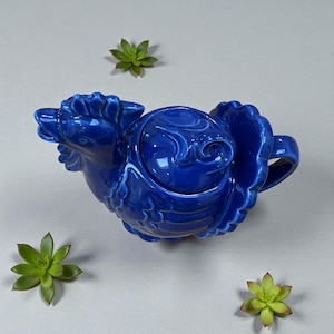 May include: A cobalt blue ceramic teapot with a rooster design. The teapot features a detailed rooster head spout, a rounded body, and a handle. The lid has a swirling design. Green succulents are placed around the teapot on a light gray surface.