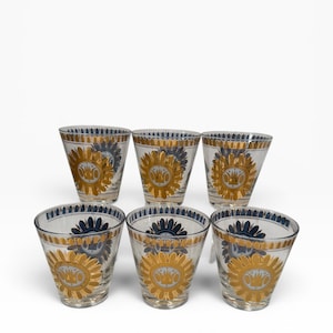May include: A set of six clear glass tumblers featuring a repeating pattern of dark blue and metallic gold sunburst motifs. Each glass has a tapered shape with a wide rim and a narrower base, decorated with gold bands along the top and bottom edges.
