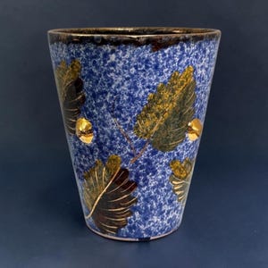 May include: A tapered ceramic tumbler featuring a speckled blue and white glaze. The exterior is decorated with several dark green and gold leaf motifs, and the rim is finished with a dark brown band. The vessel has a smooth, glossy surface.