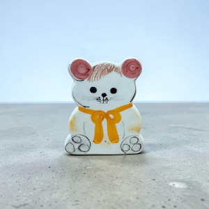 May include: A ceramic mouse figurine with a white body, brown hair, and pink ears. It has a yellow bow around its neck and black details for the eyes, nose, and feet. The figurine is sitting on a gray surface.