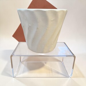 May include: A handmade, off-white ceramic cup with a textured, swirling design. The cup is displayed on a clear acrylic stand, with a geometric brown shape in the background. The cup's unique shape and color make it a decorative item.