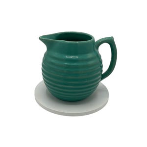 May include: A teal green ceramic pitcher with a ribbed design. The pitcher has a handle and is sitting on a white coaster.