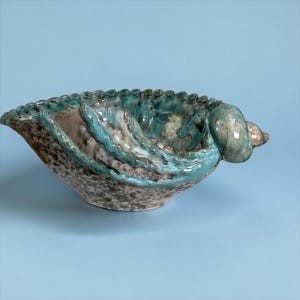 May include: A ceramic bowl shaped like a seashell with a textured, mottled gray and brown exterior. The interior features a glossy turquoise glaze with raised, wavy decorative ridges and a prominent, smooth shell-like protrusion on one side.