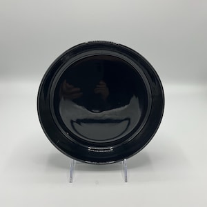 May include: A black ceramic plate with a slightly raised rim. The plate is on a clear plastic stand.