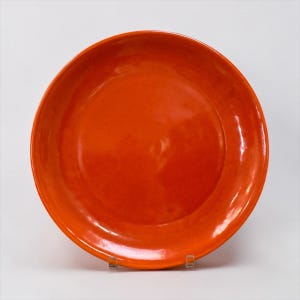 May include: A round, vibrant orange ceramic plate with a smooth, glossy finish. The plate features a wide, flat center and a slightly raised rim, showcasing a consistent, solid color throughout the entire surface of the dish.