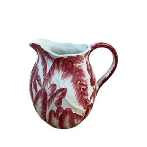 May include: A ceramic pitcher featuring a white base with a hand-painted pattern of deep red, feathery leaf-like motifs. The vessel has a rounded body, a curved handle, and a scalloped rim with a prominent pouring spout.
