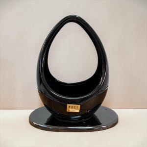 May include: A glossy black ceramic basket with a tall, arched handle and an oval base. The piece features a rectangular gold sticker on the front with the Japanese text 大宮陶芸 and the number 58 printed in black ink, centered on the lower body.
