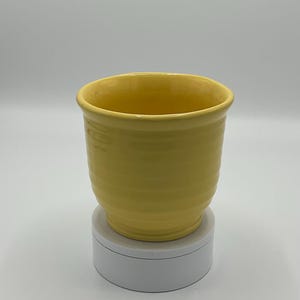 May include: A yellow ceramic planter with a smooth, rounded shape. The planter is sitting on a white pedestal.
