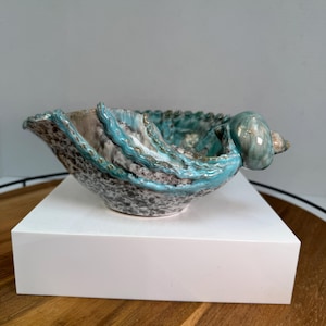May include: A ceramic bowl featuring a speckled gray and white base with raised, wavy turquoise blue accents along the rim and a sculpted, rounded shell-like protrusion extending from one side of the vessel's edge.