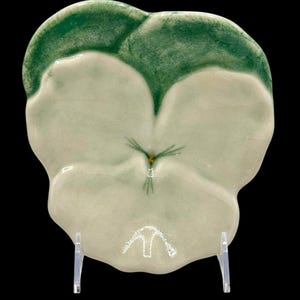 May include: A ceramic dish shaped like a pansy flower with green leaves and a white center. The dish is on a white stand.