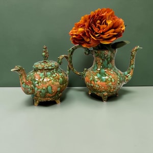 May include: A ceramic teapot with a lid and a matching pitcher, both featuring a mottled glaze of turquoise, green, and copper-orange. The pitcher contains a large cluster of vibrant orange artificial flowers with layered petals.