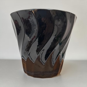 May include: A glossy black glass cup featuring a distinct spiral or twisted vertical fluted pattern. The vessel has a wide, open rim that tapers down toward a narrower base, with the bottom section showing a translucent amber-brown tint.