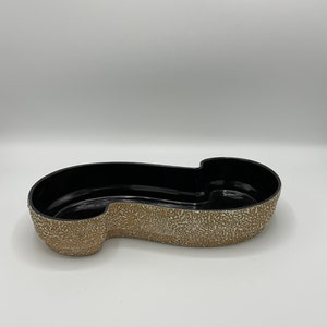 May include: A black and tan decorative dish with a unique shape. The interior is black, while the exterior has a textured, speckled tan finish. The dish has a long, curved design with a rounded section on one end.