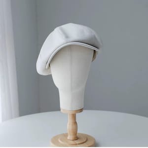 May include: A light gray newsboy cap displayed on a mannequin head. The cap has a rounded crown and a small, stiff brim. The hat is made of a soft, textured fabric. The mannequin head is mounted on a wooden stand.