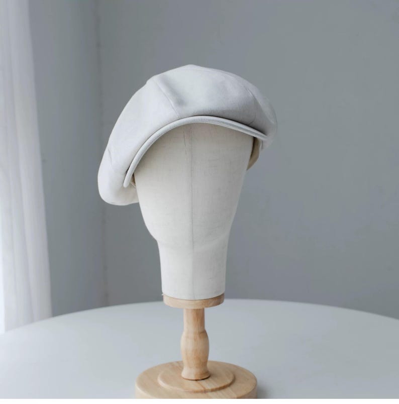 May include: A light gray newsboy cap displayed on a mannequin head. The cap has a rounded crown and a small, stiff brim. The hat is made of a soft, textured fabric. The mannequin head is mounted on a wooden stand.