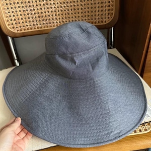 May include: A wide-brimmed, gray sun hat with a structured crown and a broad, gently curved brim. The hat is made of a textured fabric and appears to be designed for sun protection. It is resting on a chair.