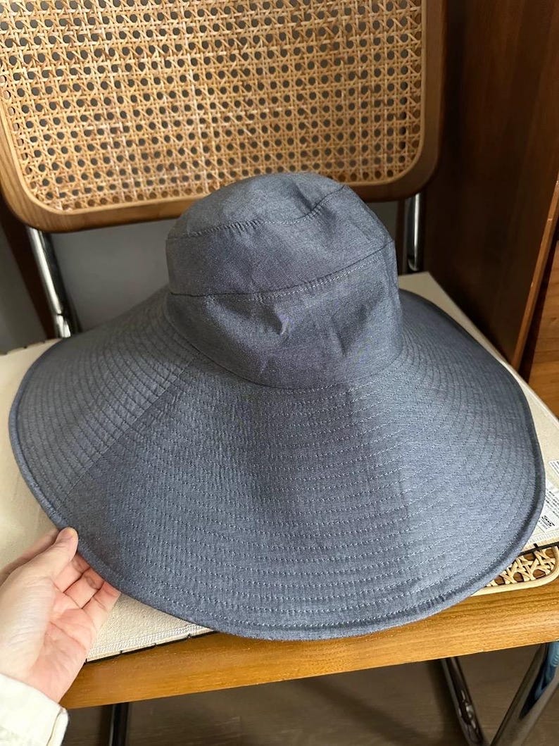 May include: A wide-brimmed, gray sun hat with a structured crown and a broad, gently curved brim. The hat is made of a textured fabric and appears to be designed for sun protection. It is resting on a chair.