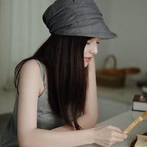 May include: A woman wearing a gray linen hat with a wide brim sits at a table and holds a piece of toast.
