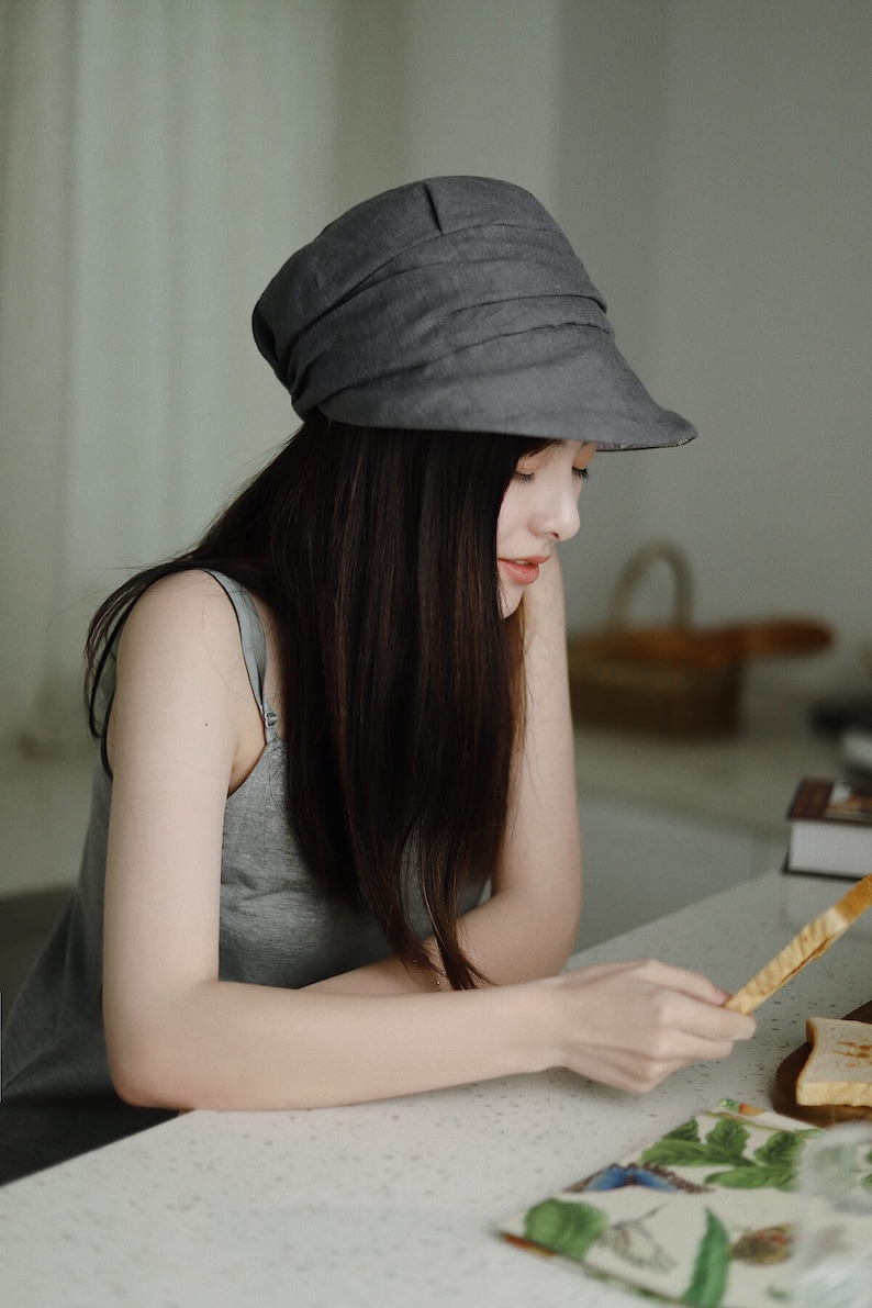 May include: A woman wearing a gray linen hat with a wide brim sits at a table and holds a piece of toast.
